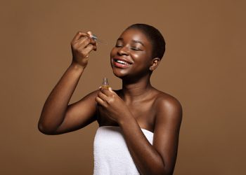 Skincare Routine. Happy Beautiful Black Female Applying Moisturizing Face Serum After Bath, Attractive African Woman With Flawless Skin Standing Wrapped In Towel Over Brown Background, Copy Space