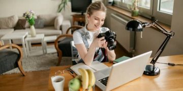 Attractive young woman photographer with blonde hair and a beautiful smile working from home, using her camera and her laptop