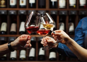 People in good mood clink glasses, celebrating together, photo focus on hands woman and man tasting degustation different types of wine, alcohol beverage to relax, bottles on shelves on background