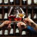 People in good mood clink glasses, celebrating together, photo focus on hands woman and man tasting degustation different types of wine, alcohol beverage to relax, bottles on shelves on background